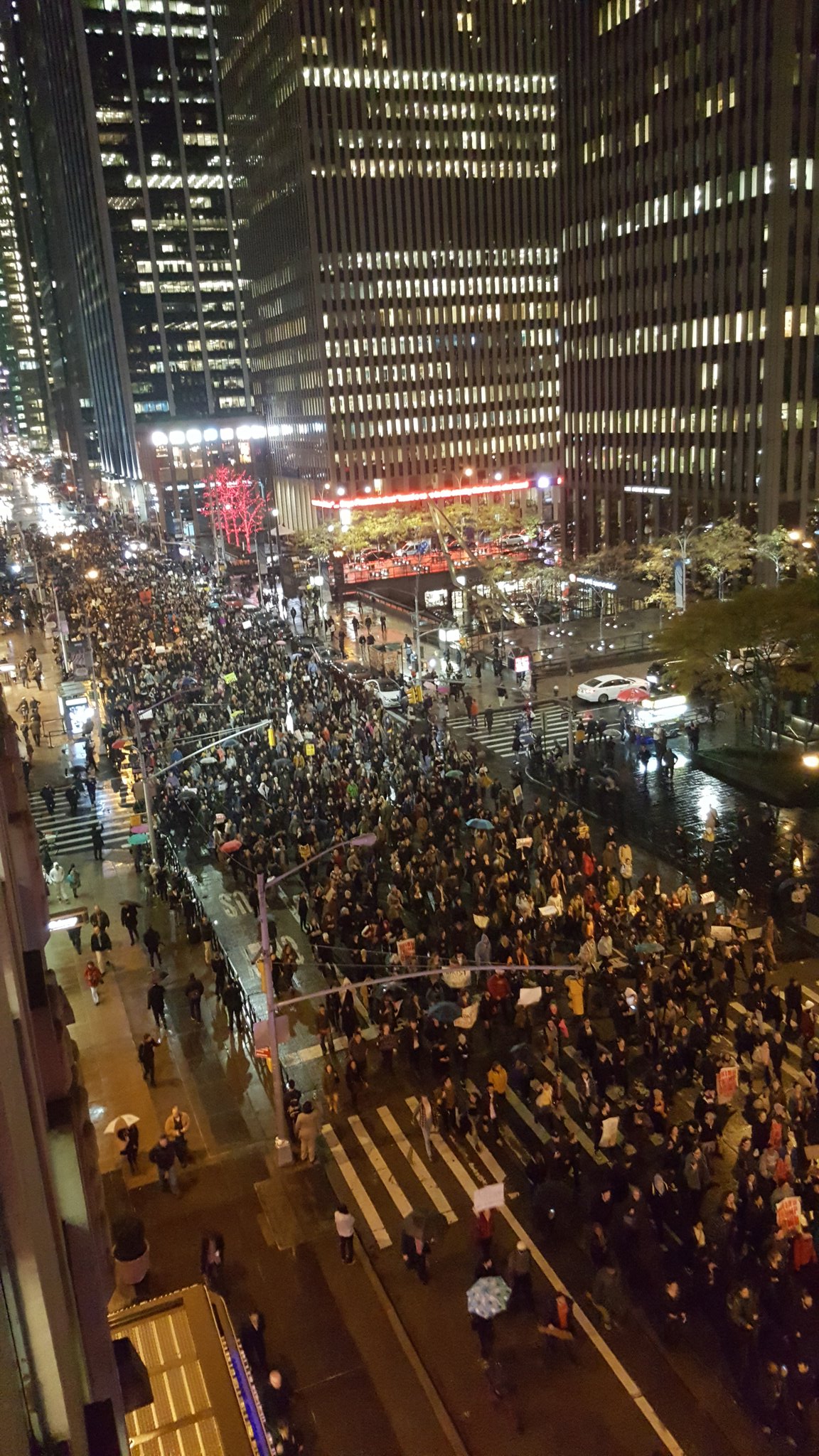 a night time shot of a large street in New York City with skyscrapers on both sides of the streets that's full of people marching